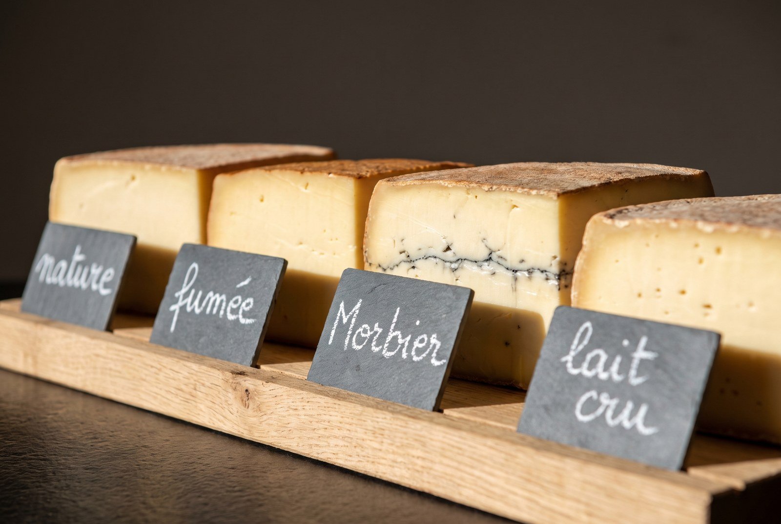 Quatre demi-meules de fromage à raclette étiquetées à la craie : nature, fumée, Morbier, lait cru